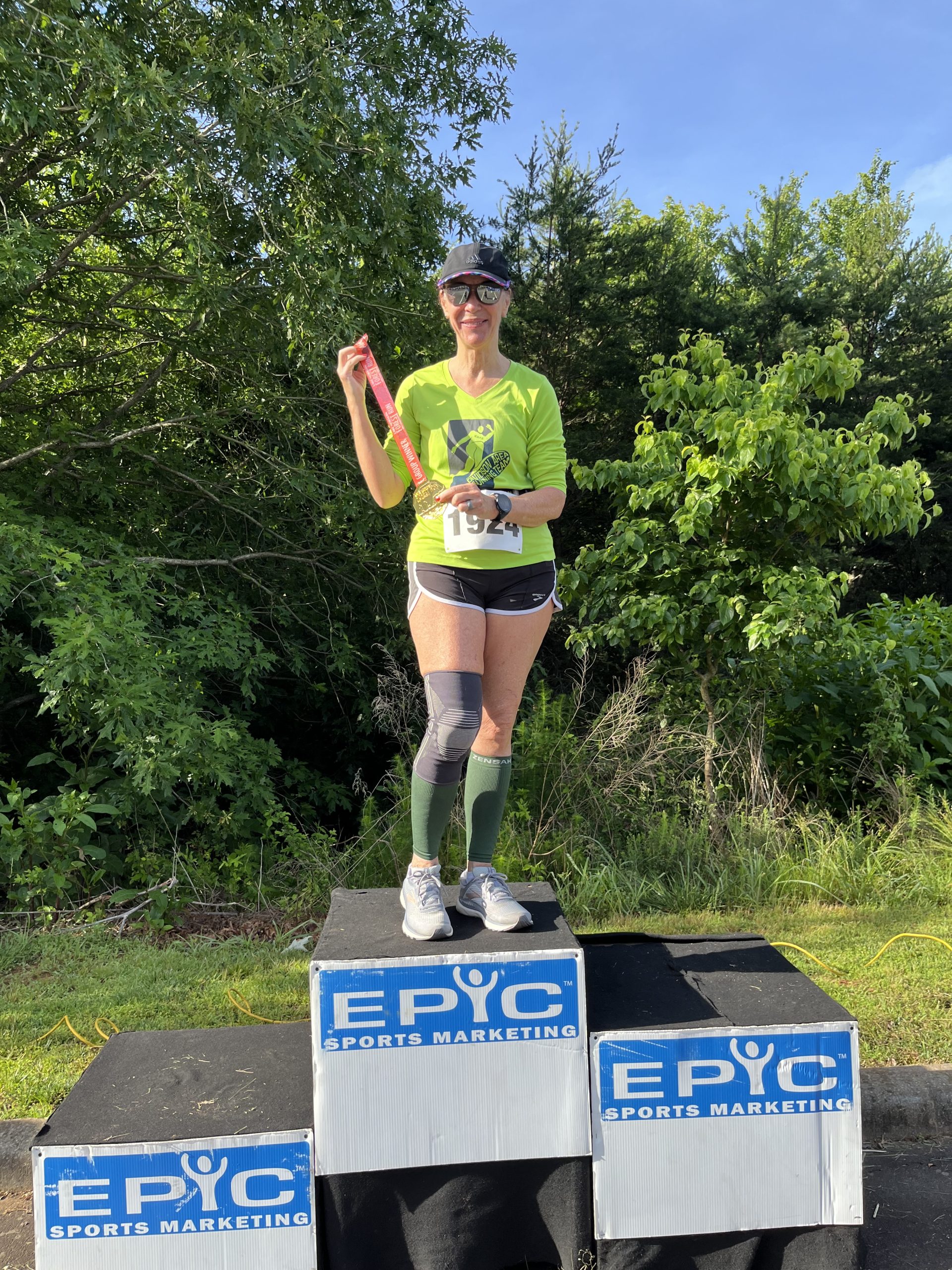 A woman in a black tank top holds a handmade poster reading 'I am never running again... oh look, a race' in front of the blue Epic Sports Marketing finish arch