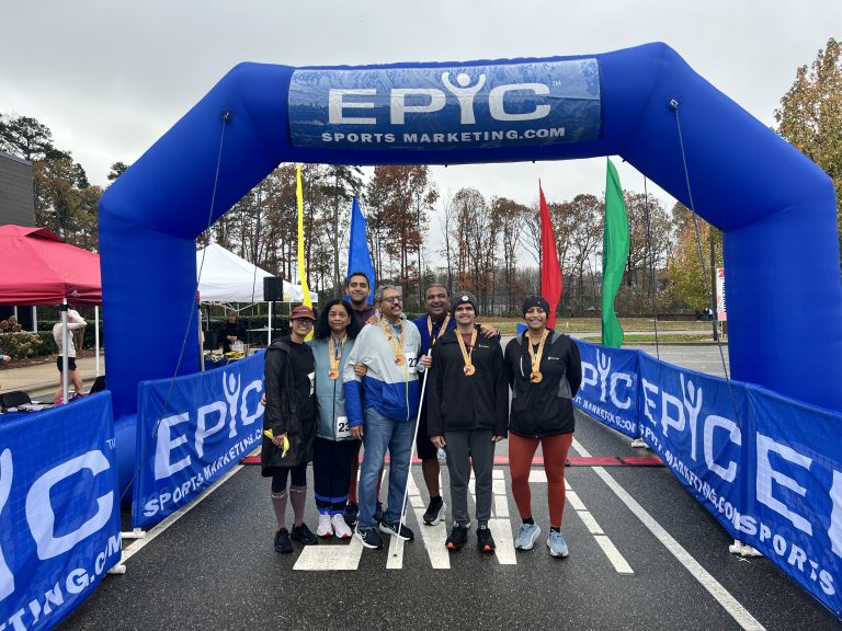 A family of seven finishers wearing medals stands together under the blue Epic Sports Marketing finish arch at the Lake Norman Turkey Trot in Cornelius, NC
