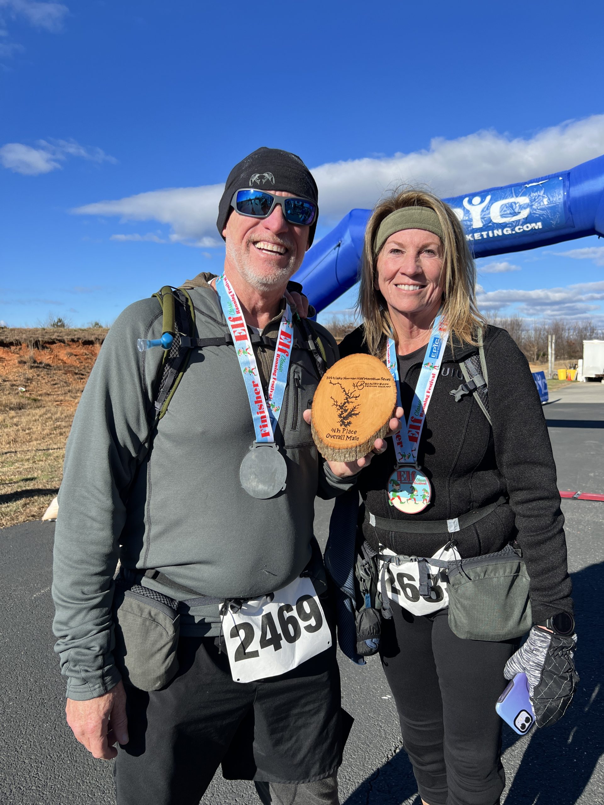A smiling man and woman wearing race bibs, finisher medals, and small hydration packs pose with a wooden award plaque beside the Epic Sports Marketing arch on a sunny winter morning